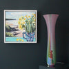 extra tall Ruth Shelley glass vase in shades of pink with central stripe d section in blues , red and greens  it is positioned next to painting of daffodils in a jug by artist Jill Hudson