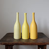A trio of Lucy Burley ceramic bottles in shades of yellow on a wooden table.