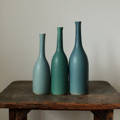 A trio of Lucy Burley ceramic bottles in shades of teal  on a wooden table.