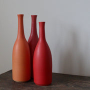 A trio of  Lucy Burley ceramic bottles in shades of red and orange sitting on a wooden table.