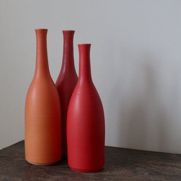 A trio of  Lucy Burley ceramic bottles in shades of red and orange sitting on a wooden table.