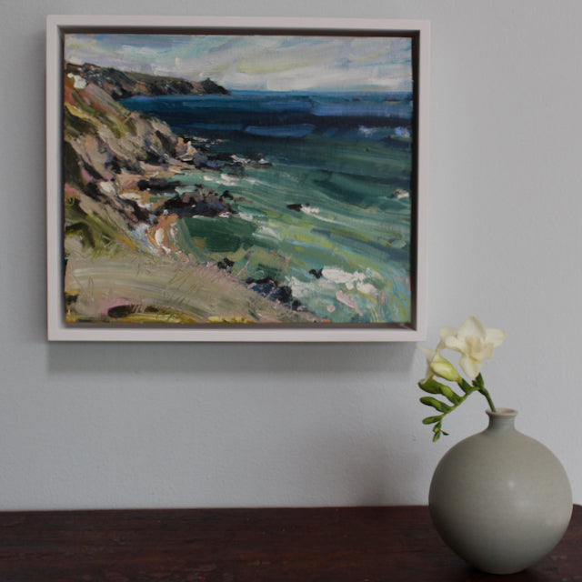 A framed painting of Rame Head in Cornwall hanging on a wall above a small green bottle on a wooden table