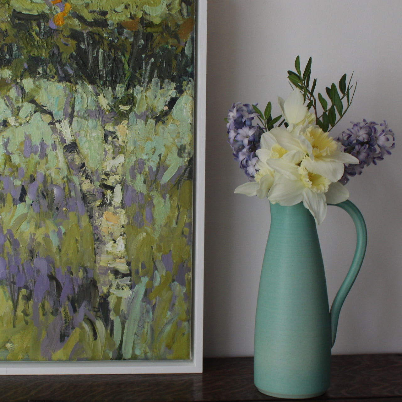 the corner of an abstract painting of trees in a meadow of purple flowers seen next to a green jug with purple flowers and pale lemon daffodils