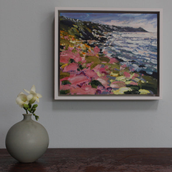 A framed painting of Rame Head in Cornwall by Jill Hudson it is hanging on a pale grey wall above a wooden table on which stands a green bottle with a freesia in it.