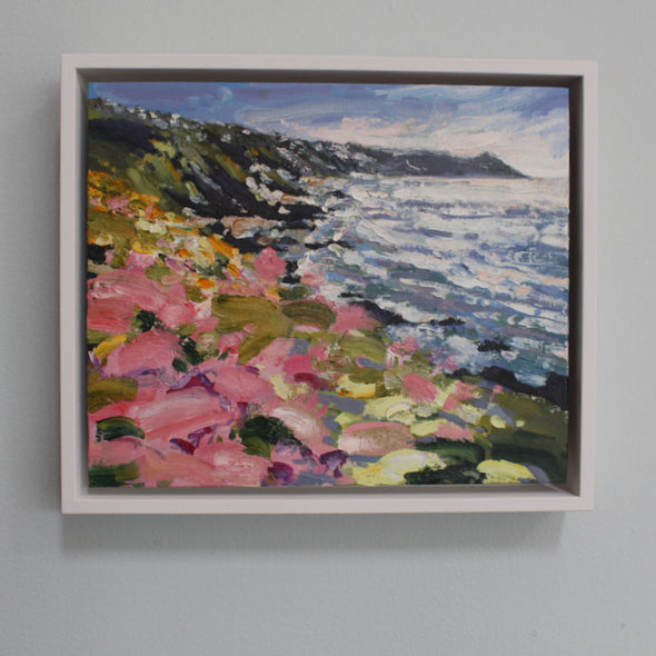 A framed painting of Rame head in Cornwall by artist Jill Hudson showing pink flowers on a cliff and a white and purple sea.