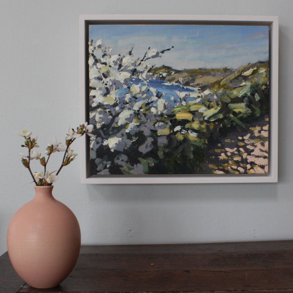 A pink vase on a table next to framed painting called April Blossom showing white blossom by Cornwall  artist Jill Hudson on a coast path in Cornwall, UK.