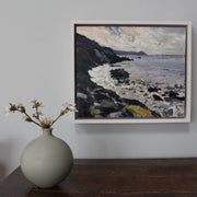 A framed painting of Rame Head in Cornwall by Jill Hudson, called From the Rocks  showing grey sea with dark cliffs  hanging  on a wall above a green bottle with white flowers in it on a wooden table