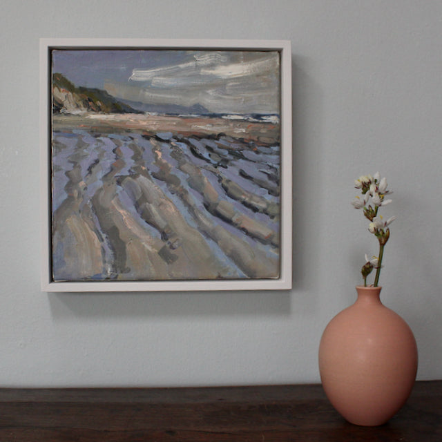 Framed painting called Tidelines,Tregantle by Jill Hudson showing tidemarks on a sandy beach on a grey day - on a table in front of the painting is a pink bottle with a white flower in it