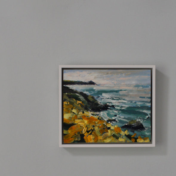 framed painting hanging called Wild Yellows hanging on a pale blue wall it depicts  a coastline with yellow flowers, is called Wild Yellows and is by Cornwall artist Jill Hudson 