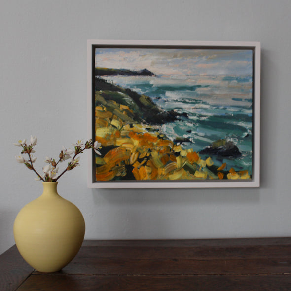 Yellow bottle with white blossom in it on a wooden table next to a framed painting of a coastline with yellow flowers, called Wild Yellows by Cornwall artist Jill Hudson 