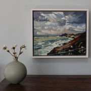 Round green bottle with blossom in it on a table next to a framed painting showing cloudy skies and the coastline by Cornwall artist Jill Hudson.