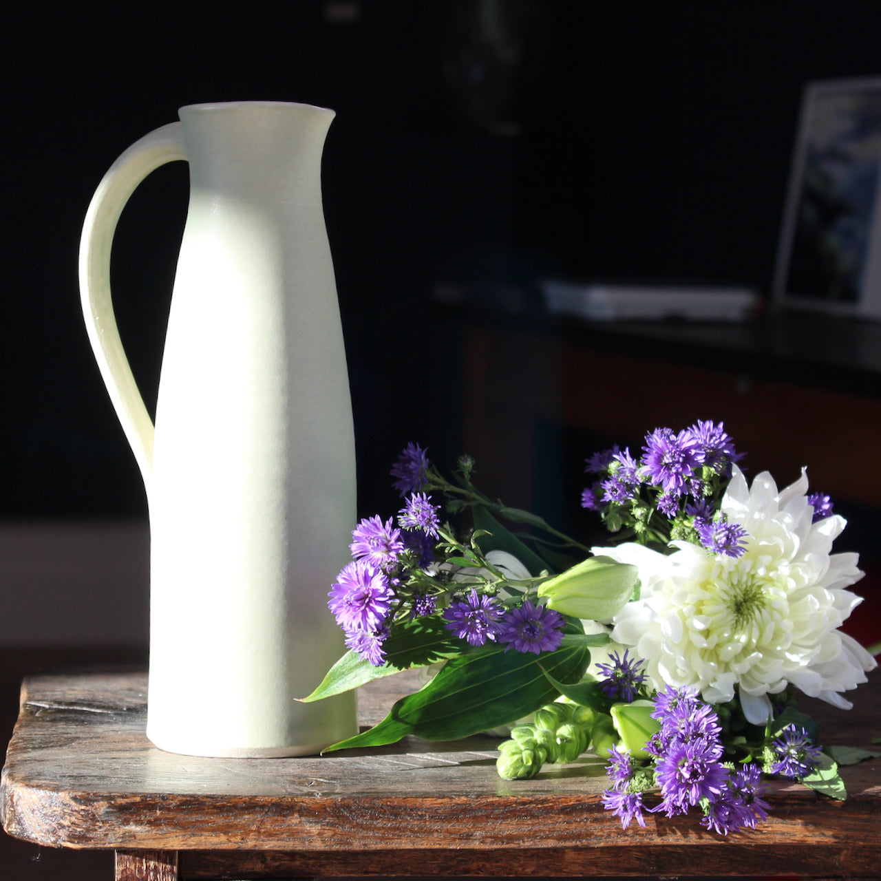 pale olive ceramic jug by UK ceramicist Lucy Burley with a bunch of white and purple flowers lying beside it 