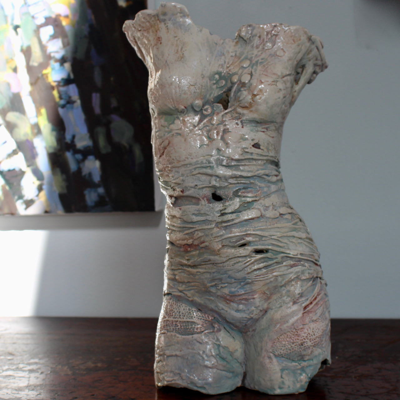 a textured ceramic torso made by Pauline Lee standing on a wooden table with the edge of a painting in the background