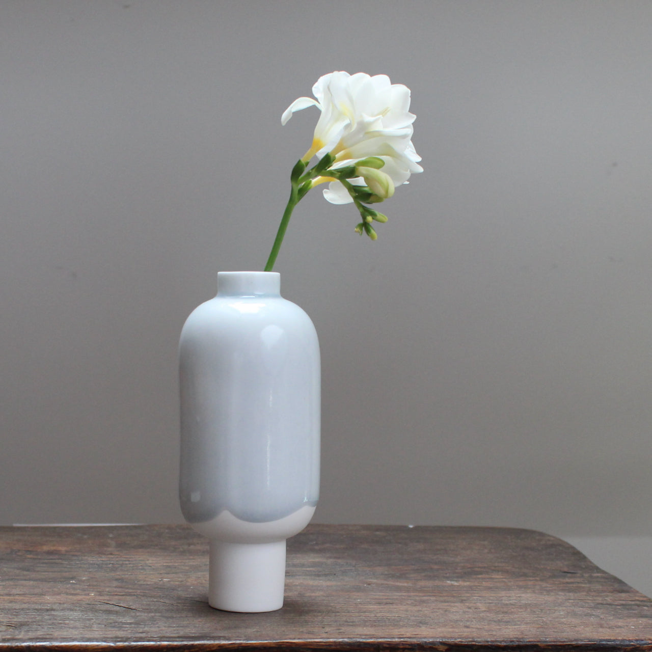 Small pale blue and white ceramic vessel with a single white freesia stem in it standing on a wooden table 