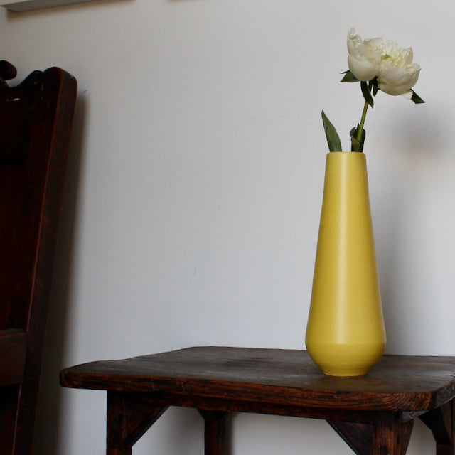 tall yellow vase by Lucy Burley it has a white flower in it and it's sitting on a wooden table