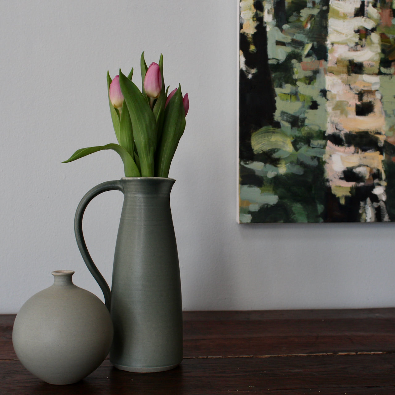 pale green bottle and a darker green jug with tulips in it on a wooden table with corner or a painting visible 