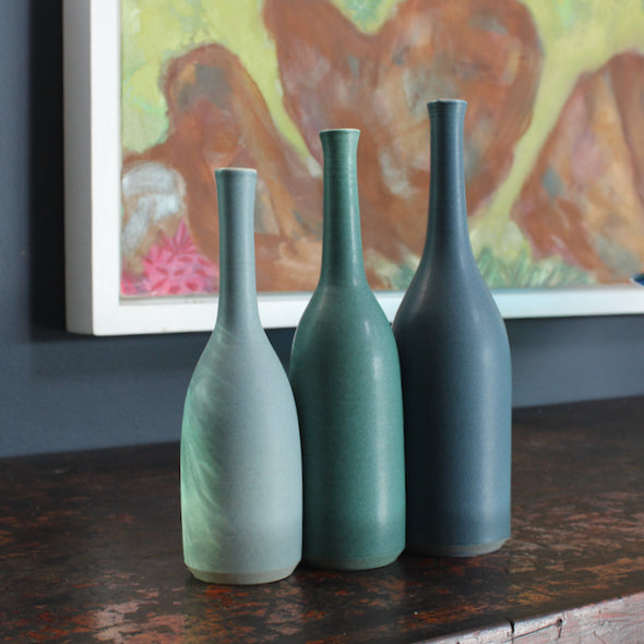 A trio of Lucy Burley ceramic bottles in shades of teal  on a wooden table in front of a painting.