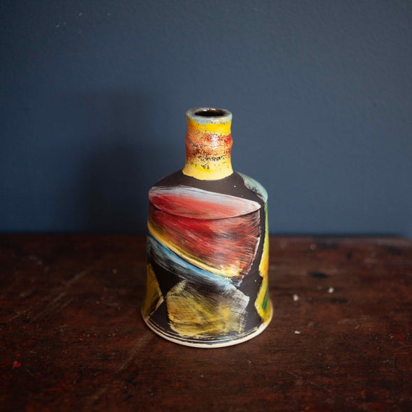 A brightly coloured ceramic bottle by John Pollex on a wooden table with  a blue wall behind