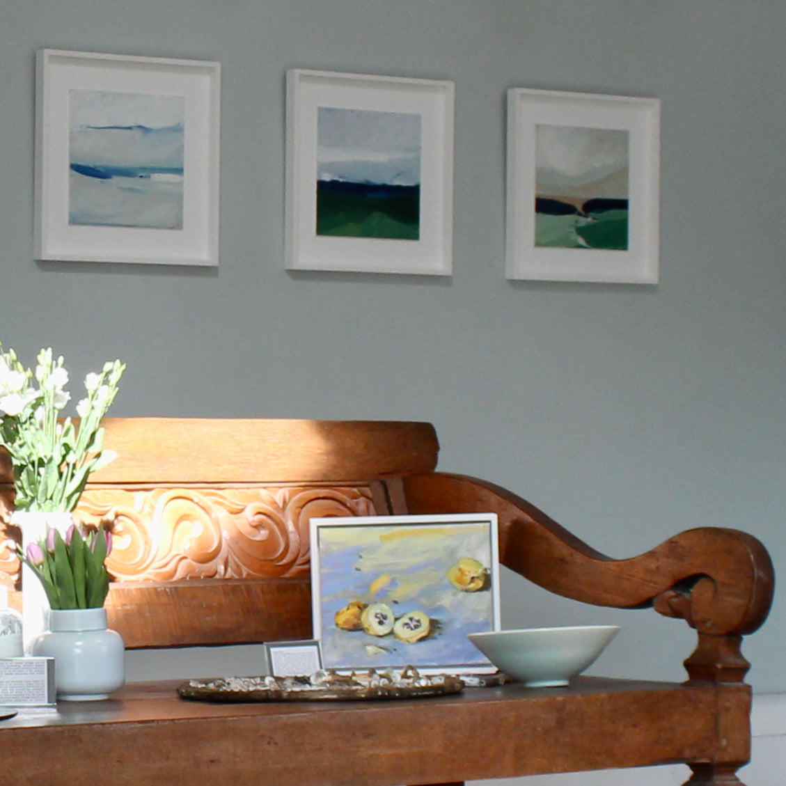 Three paintings of abstract landscapes on a gallery wall above a wooden bench.