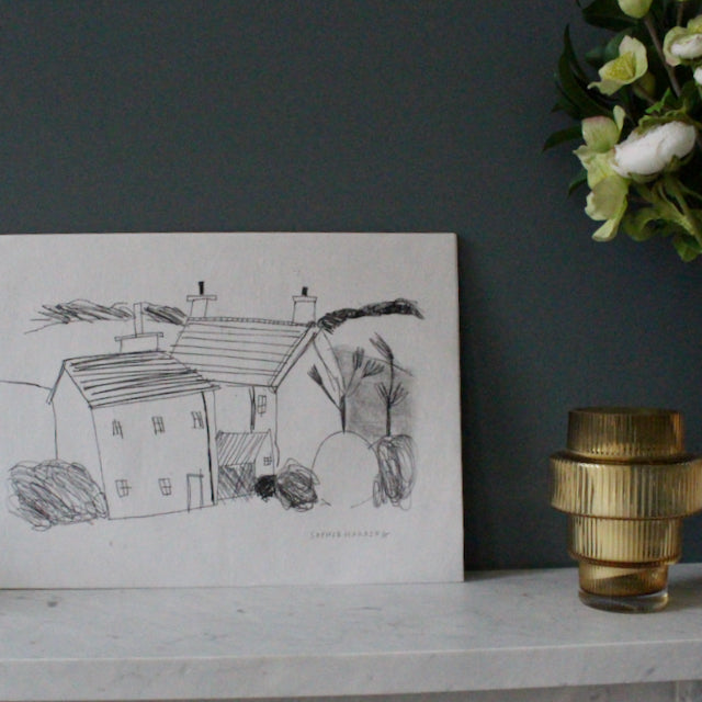 a drawing in black ink on white paper of Cornish cottages by artist Sophie Harding it is leaning against a grey wall resting on a marble mantlepieces with a vase of flowers beside it.