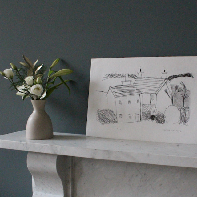 a drawing in black ink on white paper of Cornish cottages by artist Sophie Harding it is leaning against a grey wall resting on a marble mantlepieces with a vase of flowers next to it