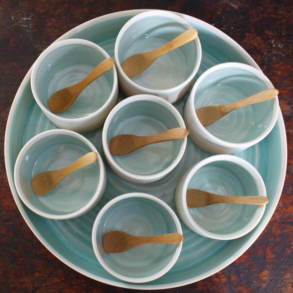 Seven small bowls with turquoise interiors and wooden spoons set inside larger serving dish
