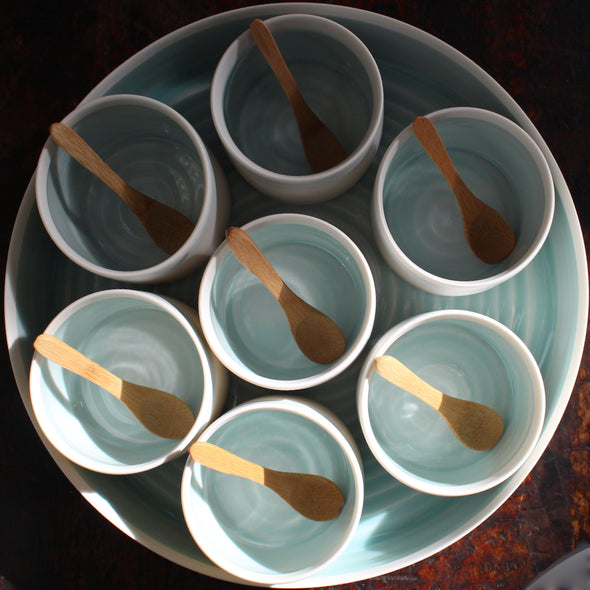 Seven small bowls with turquoise interiors and wooden spoons set inside a larger serving dish