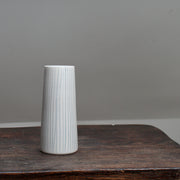 A narrow ceramic white stem vase with vertical blue lines on a wooden table .