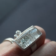 Silver ring featuring large rectangular quartz stone with small diamond and small silver ball to the side shown on a finger