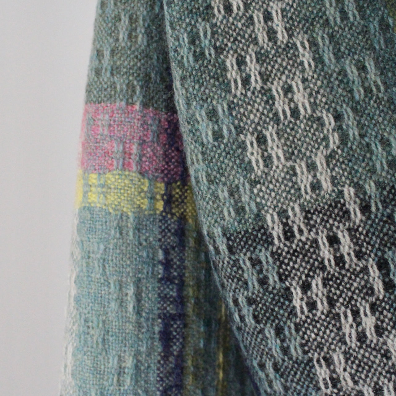 close up of a fringed handwoven scarf by UK textile artist Teresa Dunne it's in shades of green, blue, yellow and pink 