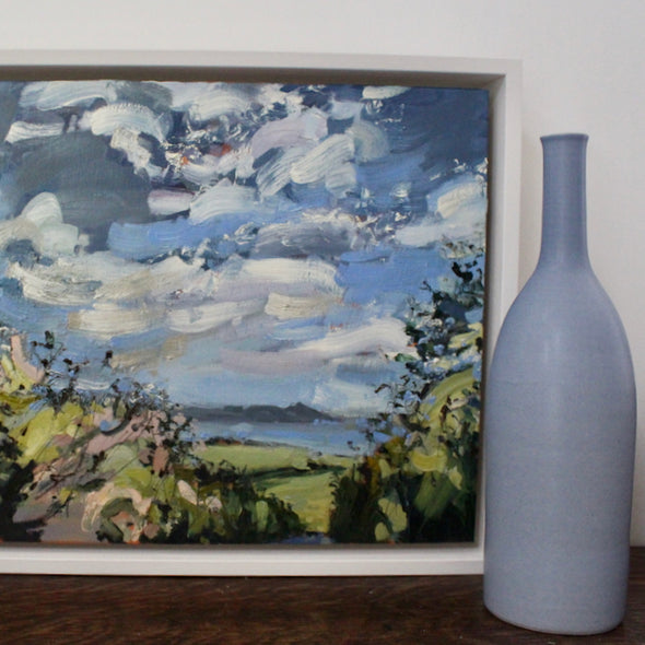 painting of a view of Rame Head in south east Cornwall with pink and green flowers in the foreground and white clouds in the sky by Cornwall artist Jill Hudson  it is next to a blue ceramic bottle by Lucy Burley.