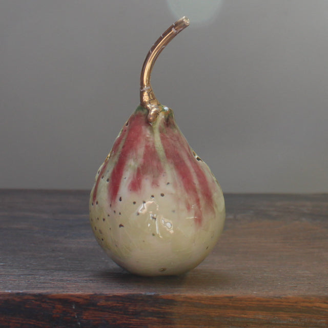 A cream and pink ceramic pear with glaze details and a gold stalk by ceramicist Remon Jephcott.