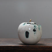 sitting on a wooden table, a large ceramic white apple with a gold stalk by Remon Jephcott.