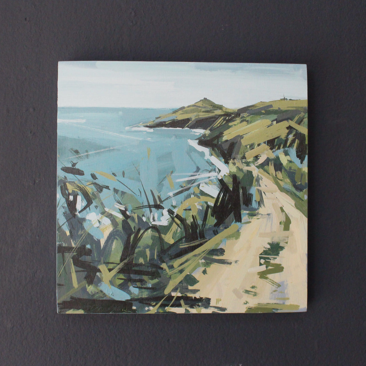 An Imogen Bone landscape painting of Rame Head in south east Cornwall showing headland in greens and browns and sea in blue it is hanging on a dark blue wall