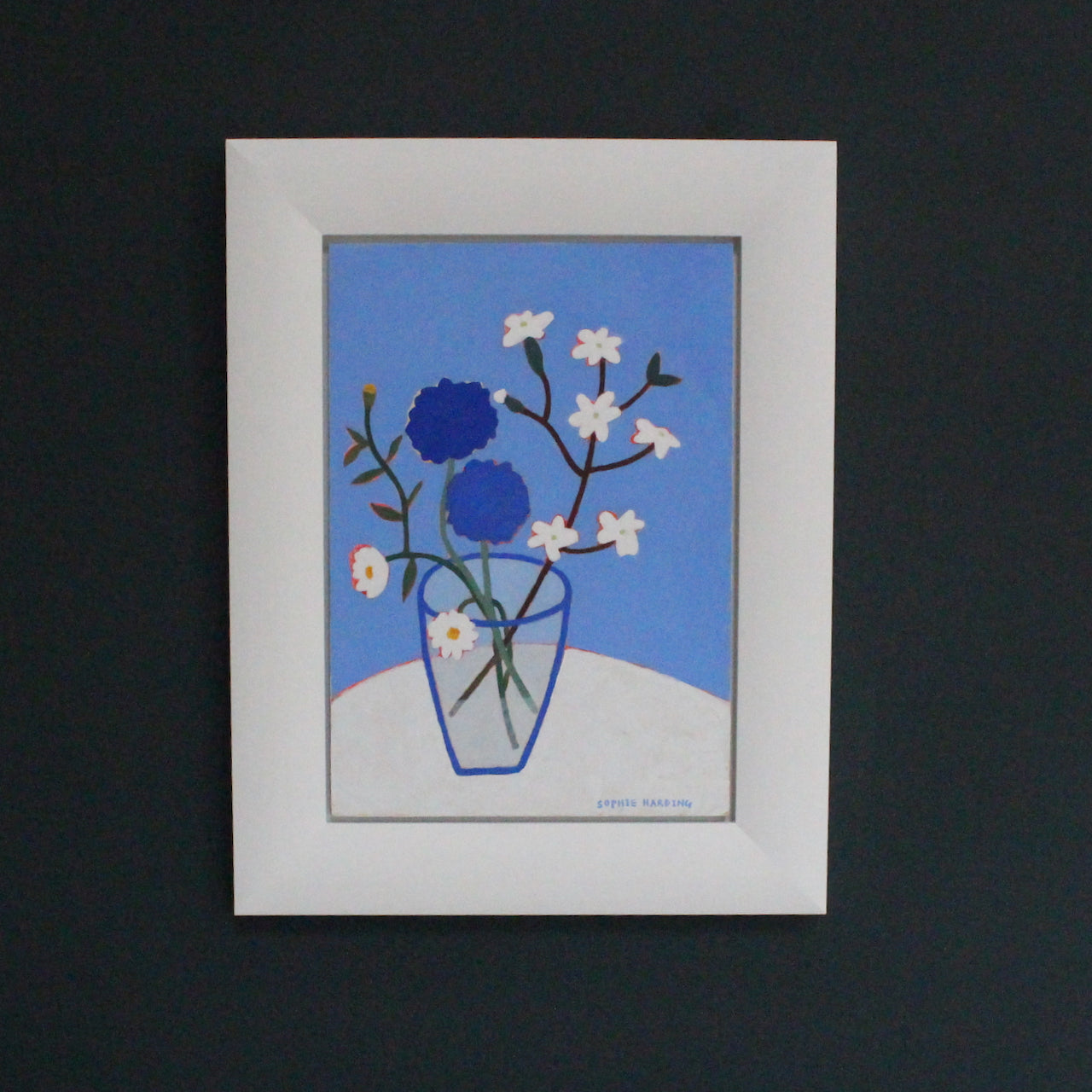 a framed Sophie Harding painting of painting of white and blue cornflowers in a glass vase on a white table cloth 