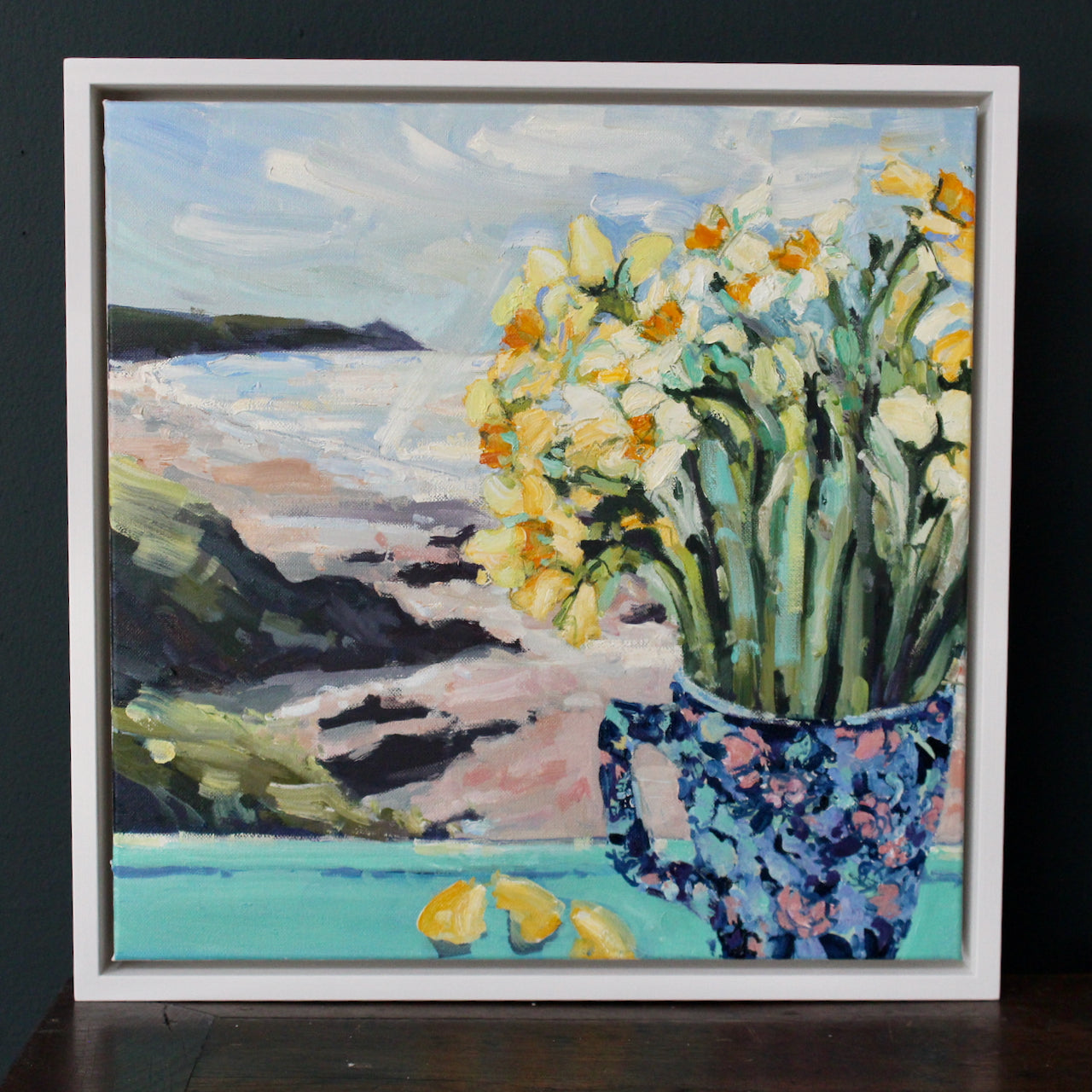 a framed Jill Hudson oil painting of yellow daffodils in a blue patterned jug on a windowsill looking out to Rame Head peninsula in Cornwall 