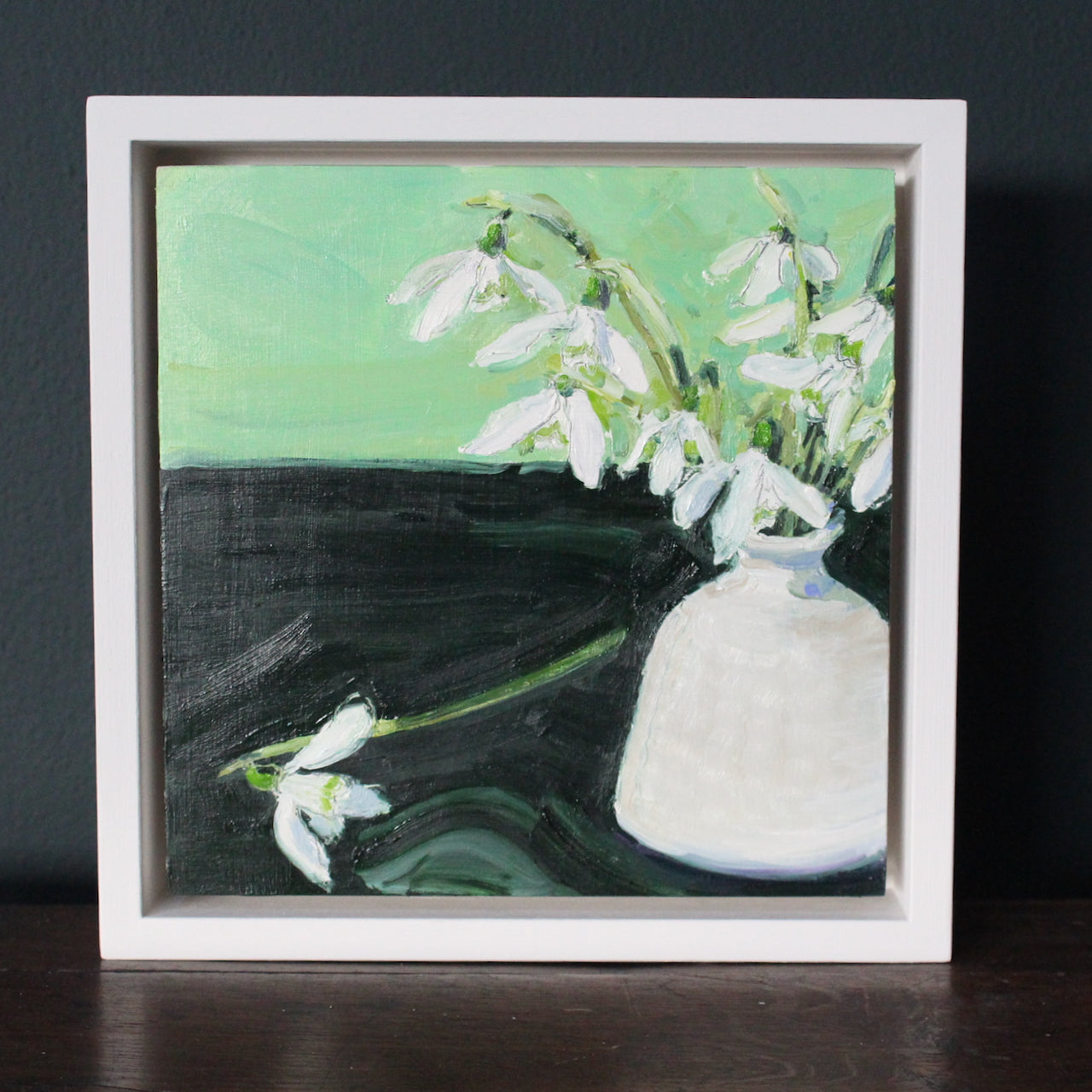 framed Jill Hudson oil painting of snowdrops in a small white vase against a green background.