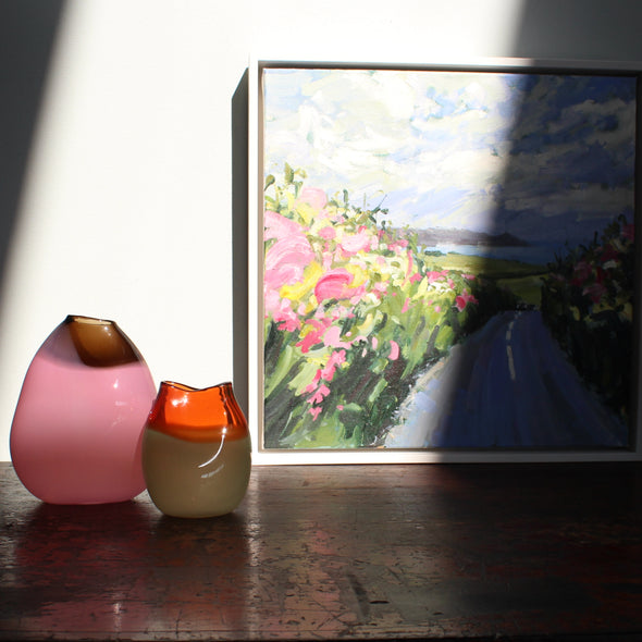 a painting by Cornwall artist Jill Hudson of a colourful hedgerow and the view towards Rame Head in south Cornwall, the painting is positioned next to two glass vessels one pink and one green by glass artist Michele Oberdieck