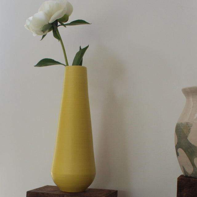 A tall yellow vase by Lucy Burley it has a white flower in it and it's sitting on a wooden table next to another pottery vase which is just seen